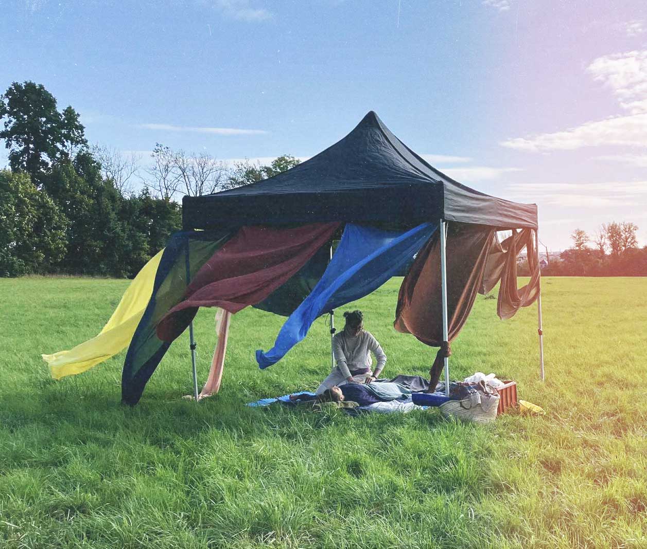 Person sitting under a black canopy tent with colorful fabric panels, massaging another person lying on a blanket in a grassy field.